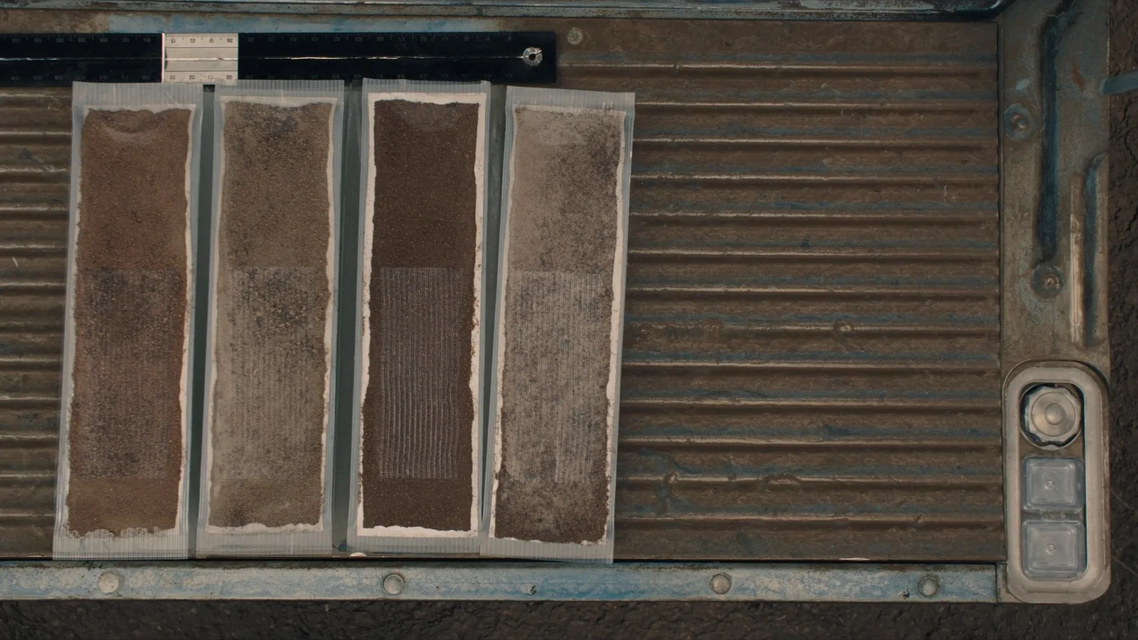 Four sleeved soil samples laid side by side on a steel truck tailgate with a folding rule across the top of them, the upper part of each sample a red brown clay and the lower part a paler grey.