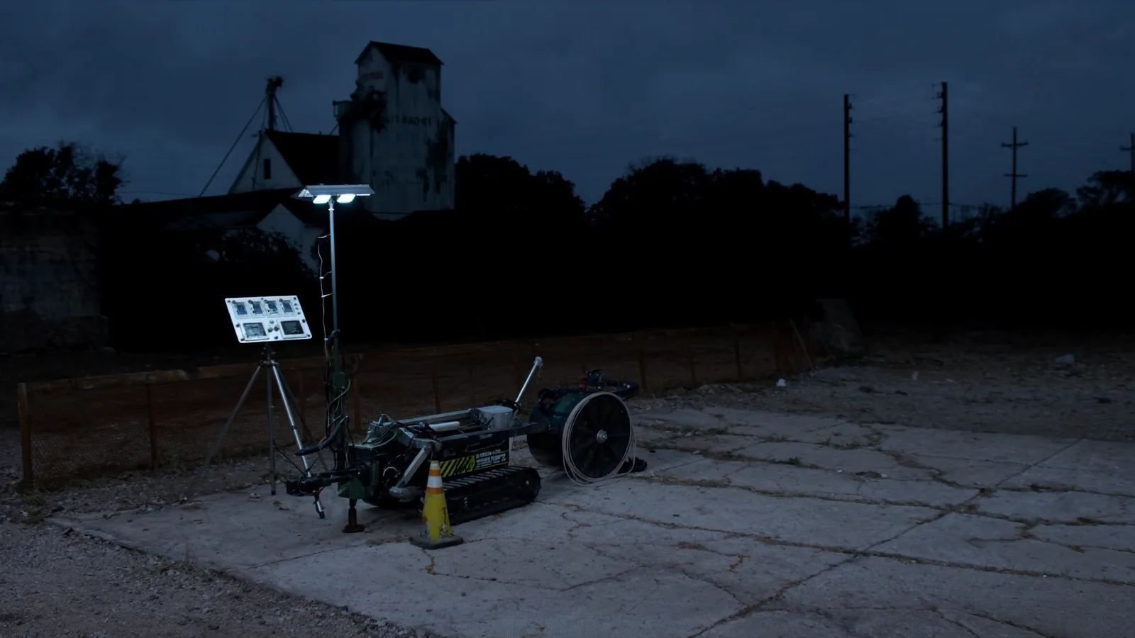 A track mounted direct push drilling rig set up under a work light on a cracked concrete lot at dusk, with a grain elevator on the skyline behind it.